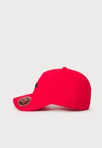 Rød cap lavet af bomuld med en buet skygge, der har et sort logo på fronten og et rundt sort mærke på siden. Struktureret design.