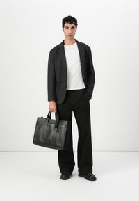 Svart lær tote-veske med doble topphåndtak, strukturert form, preget logo og avtagbar merkelapp, sammen med en grå blazer og svarte bukser.