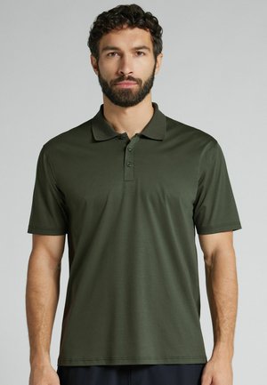 Muž s vousem v tmavě zelené polokošili s krátkým rukávem stojí před jednoduchým světle šedým pozadím.