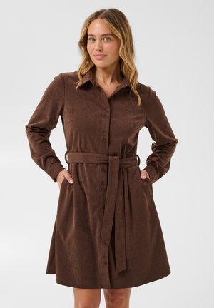 Femme aux cheveux châtain clair portant une robe-chemise en velours côtelé marron à manches longues avec des boutons et une ceinture nouée à la taille, les mains dans les poches.