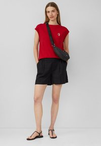 Rode t-shirt met korte mouwen met een witte grafische afbeelding, gecombineerd met zwarte shorts en zwarte sandalen. Een gestructureerde zwarte crossbody tas wordt gedragen.
