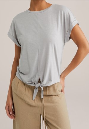 Femme portant un haut gris clair à manches courtes avec un nœud devant et un pantalon beige taille haute, debout avec une main sur la hanche.