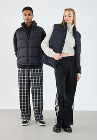 Veste doudoune noire avec capuche, associée à un col roulé en tricot gris clair. Pantalons de survêtement noirs avec rayures blanches et pantalons à carreaux. Baskets noires.