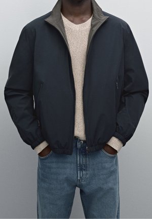 Mann trägt eine schwarze Zip-Up-Jacke über einem beige gestrickten Pullover, die Hände in den Taschen hellblauer Jeans, vor neutralem Hintergrund.