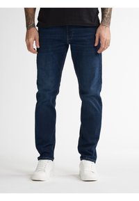 Tmavě modré jeansy z denimu s úzkým střihem, viditelnými švy a rovným střihem nohavic, doplněné o bílé tenisky.