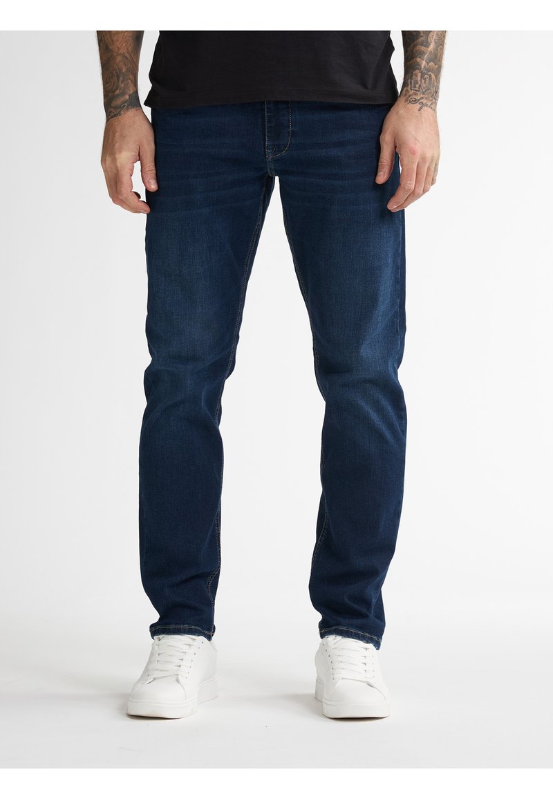 Tmavě modré jeansy z denimu s úzkým střihem, viditelnými švy a rovným střihem nohavic, doplněné o bílé tenisky.