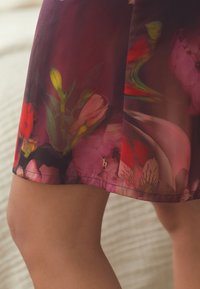 Gėlių raštuota sijonas violetinių ir rožinių atspalvių, pagamintas iš lengvos medžiagos, su auksiniu akcentuotu logotipu apatinėje dalyje ir lygiu paviršiumi.