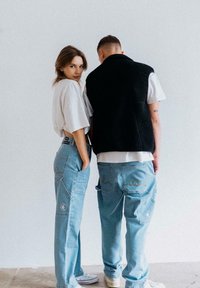 Hellblaue Jeans mit lockerer Passform, mit Taschenakzenten und Stickerei. Eine Person trägt ein weißes, übergroßes T-Shirt, die andere eine schwarze Weste.