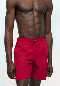 Άνω σώμα άνδρα με σκουρόχρωμο δέρμα που φορά φωτεινά κόκκινα μαγιό με κορδόνι, στέκεται μπροστά σε μια απλή ανοιχτή επιφάνεια.