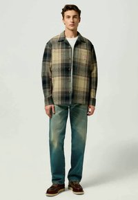 Camisa de botones a cuadros en tonalidades de beige, negro y azul marino; combinada con jeans azul desgastado y zapatos marrones. Presenta un pequeño parche con logo.