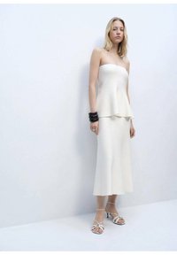 Bílý top bez ramínek s peplum střihem spárovaný s odpovídající midi sukní, vyrobený z hladkého materiálu. Modelka nosí černé náramky a bílé sandály na podpatku.