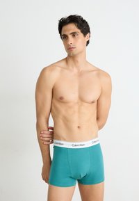 Teal bomull boksershorts med en hvit midjebånd som har "Calvin Klein" logo, tettsittende design og glatt tekstur. Modell vist fra siden.