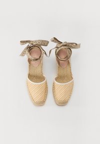 Béžové espadrilky s tkaným svrškem, bílým lemem a hnědými vzorovanými kotníkovými stuhami. Podešev je vyrobena z pleteného jutového materiálu pro texturu.