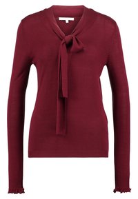 Vínový pletený svetr s vázáním u krku, dlouhými rukávy a žebrovanými manžetami se volánem. Měkká textura s upnutým střihem.