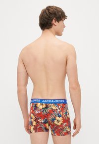 Red floral boxer shorts feature a blue branded waistband. The design includes vibrant tropical patterns with leaves and flowers.