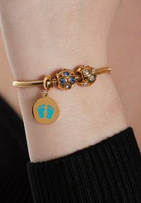 Gold bracelet with two decorative beads, one featuring blue stones and the other clear stones. A charm with teal footprints hangs from the design.