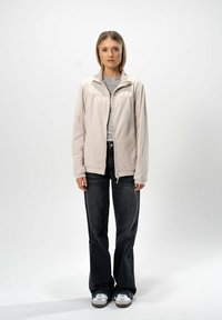 Vaaleanbeige vetoketjullahka korkealla kauluksella, yhdistettynä harmaaseen T-paitaan ja tummiin farkkuihin. Malli seisoo yksinkertaisella taustalla.