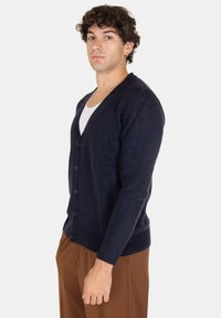 Giovane uomo con capelli ricci, che indossa un cardigan blu scuro con bottoni sopra una camicia bianca e pantaloni marroni, in piedi di fronte a uno sfondo neutro.