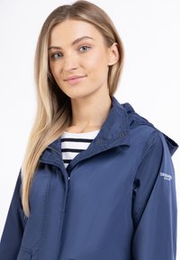 Giacca impermeabile blu navy con chiusura a zip, cappuccio regolabile e tasche, indossata sopra una camicia a righe. Presenta un logo sulla manica.