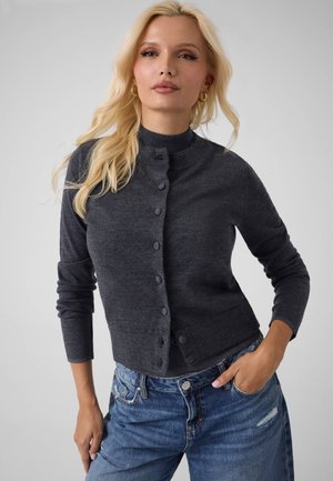 Femme blonde portant un cardigan gris foncé boutonné et un jean bleu, une main dans la poche, devant un fond uni.