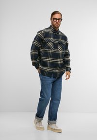 Granatowa flanela w żółto-kremową kratę, z dwoma kieszeniami na piersi, mankietami na guziki, zestawiona z luźnymi niebieskimi dżinsami i beżowymi sneakersami.