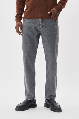 Grijze denim jeans met een rechte pasvorm, raw hem en een lichte vervaging. Gecombineerd met zwarte instappers. Een trui in een bruine tint completeert de look.
