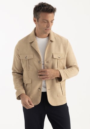 Homme portant une veste beige avec poches à rabat sur une chemise blanche et un pantalon noir, ajustant le devant de la veste, sur fond blanc uni.