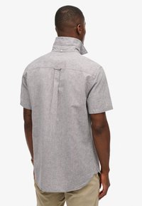 Camicia grigia a maniche corte con colletto, caratterizzata da una texture sottile, una fessura sul retro e un dettaglio a bottoni sul colletto. Indossata con pantaloni color khaki.