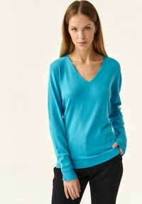 Pulover turcoaz cu decolteu în V, realizat dintr-un material moale, având mâneci lungi și manșete din bumbac cu un design ajustat. Asortat cu pantaloni negri.