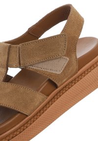 Suede tan sandal with cut-out design, featuring brown rubber sole and braided detailing along the side, comfortable footbed, and adjustable straps.