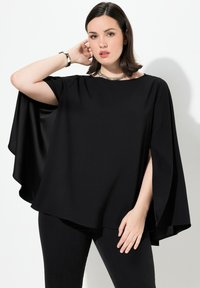 Top nero in stile cappotto con maniche corte, realizzato in tessuto morbido e dal fitting rilassato. Indossato con pantaloni neri, mostra dettagli sulle braccia e sulla vita.