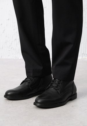 Czarne skórzane buty o teksturowanym wykończeniu, z sznurowadłami, sięgające kostki, z płaską gumową podeszwą, w zestawie z czarnymi spodniami.