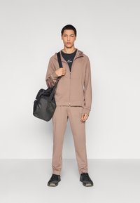 Φωτεινό καφέ φούτερ με φερμουάρ και αντίστοιχα παντελόνια φόρμας, φορεμένα πάνω από μαύρο T-shirt, συνδυασμένα με μαύρα αθλητικά παπούτσια και μαύρη τσάντα.