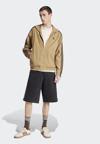 Giacca zip-up color sabbia con maniche a righe nere, abbinata a pantaloni corti in denim scuro. Una maglietta bianca e sneaker color sabbia con dettagli marroni completano l'outfit.
