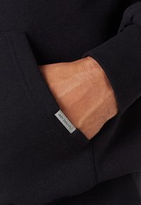 Zwarte sweatshirts met een kangoeroezak en geribde manchetten, voorzien van een klein metalen logotagje aan de rand van de zak. Zacht stoftextuur.