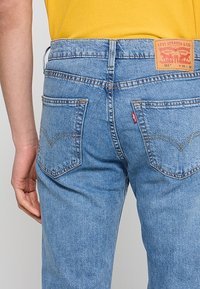 Personne portant un jean Levi's 511 bleu clair avec les poches arrière visibles et une chemise jaune rentrée.
