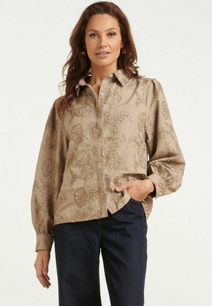 Vrouw draagt een beige longsleeve met bloemrijke borduurwerk en donkerblauwe spijkerbroek, staand met één hand in haar zak.