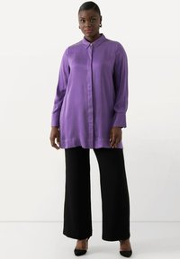 Blusa lunga viola con colletto con bottoni, abbinata a pantaloni larghi neri e tacchi neri. Tessuto setoso, design minimale.