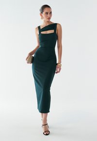 Zielona sukienka midi z jednym ramieniem, z kwadratowym wycięciem i teksturowaną tkaniną. Uzupełniona złotą kopertówką i czarnymi sandałami na cienkich paskach.
