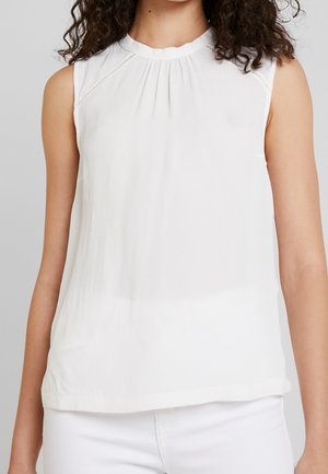 Vrouw draagt een mouwloze witte blouse met subtiele plooien bij de halslijn en bijpassende witte broek tegen een lichtgrijze achtergrond.