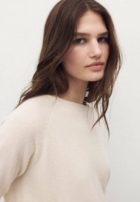 Un pull en tricot texturé beige clair avec un col rond. Le modèle a les cheveux bruns à mi-longueur et une expression faciale neutre.