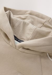 Sweat à capuche beige avec texte visible sur l'étiquette intérieure "BAKER" et gros plan sur un tissu doux et texturé.