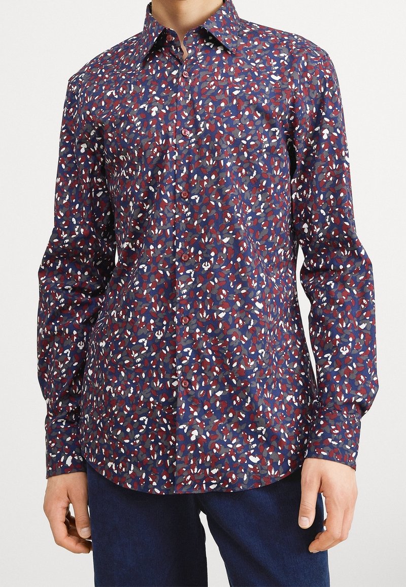 Chemise à manches longues boutonnée avec un motif abstrait multicolore de feuilles en rouge, blanc et bleu portée par une personne debout devant un fond uni.