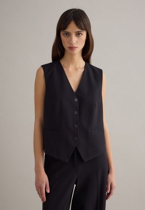 Junge Frau mit glattem braunem Haar, die eine ärmellose schwarze Weste mit Knöpfen und passende schwarze Hose vor einem beigen Hintergrund trägt.