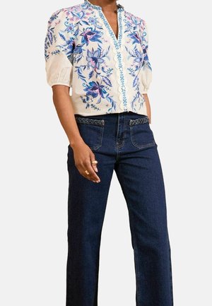 Femme portant un chemisier blanc avec une broderie florale bleue et un jean bleu foncé avec un détail tressé sur les poches.