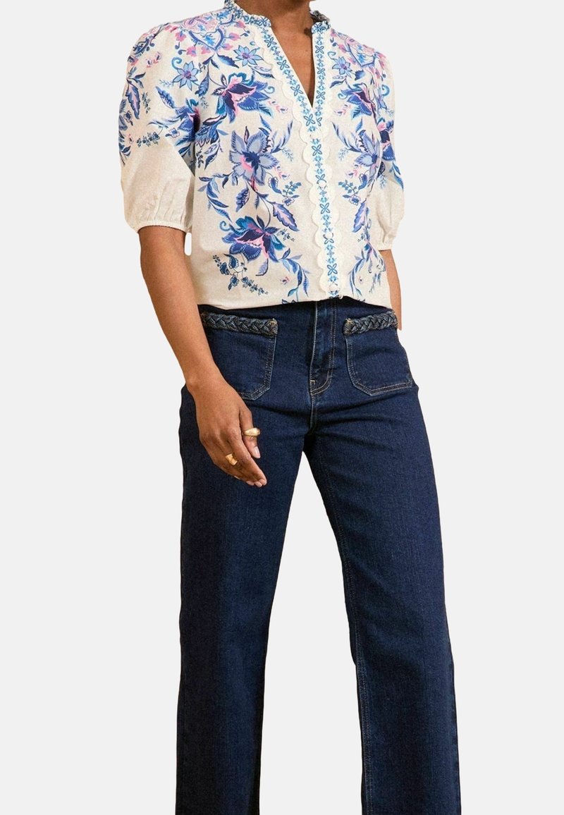 Femme portant un chemisier blanc avec une broderie florale bleue et un jean bleu foncé avec un détail tressé sur les poches.