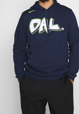 Homme portant un sweat à capuche bleu marine avec le texte "DAL" et le logo des Dallas Mavericks, associé à un pantalon de jogging noir Nike, les mains dans les poches.