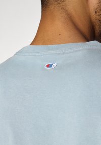 Lys blå bomulls T-skjorte med rund hals, med en liten brodert logo i rødt og blått på baksiden nær nakken.