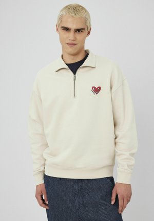 Sweatshirt zippé crème avec des poignets côtelés, présentant un design de cœur rouge avec des yeux et un sourire. Porté par-dessus une chemise navy, tissu texturé.