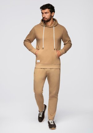 Mann mit Bart, der einen beigen Hoodie und passende Jogginghose trägt und mit Händen in den Taschen des Hoodies vor einfachem Hintergrund läuft.